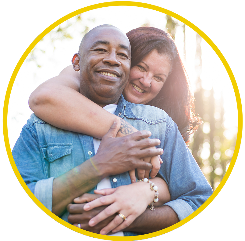 A woman stands behind a man and puts her arm around him as they stand in a park and smile.